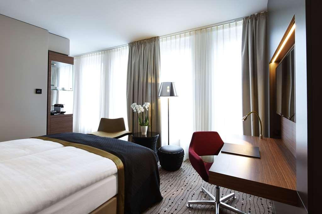 Steigenberger Hotel Am Kanzleramt Берлін Номер фото The photo shows a modern hotel room featuring a bed with a white comforter and a dark accent blanket. On one side of the room, there are large windows with sheer curtains allowing natural light to enter. A small desk with a lamp is set up near the wi