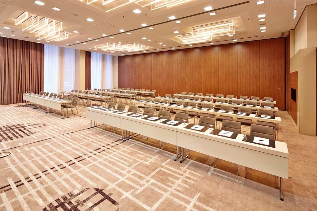 Steigenberger Hotel Am Kanzleramt Берлін Зручності фото The photo shows a modern conference room set up for a meeting or seminar. There are multiple rows of long, white tables arranged in a rectangular layout, each equipped with chairs. On the tables, there are items like notepads and pens, indicating a p
