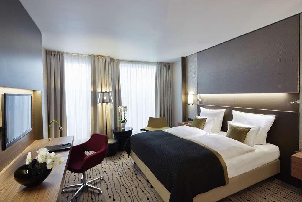 Steigenberger Hotel Am Kanzleramt Берлін Номер фото The photo shows a modern hotel room with a minimalist design. It features a large double bed with white linens and a dark throw blanket. On either side of the bed, there are small lamps. There is a desk with a chair, and a flat-screen TV mounted on t