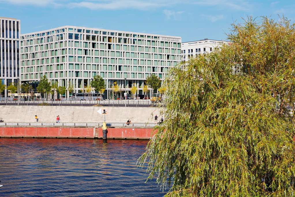 Steigenberger Hotel Am Kanzleramt Берлін Екстер'єр фото The photo shows a modern building with a distinctive geometric design situated near a body of water. In the foreground, there is a river, and a green willow tree or similar vegetation is visible. The area appears to be a waterfront promenade, with pe