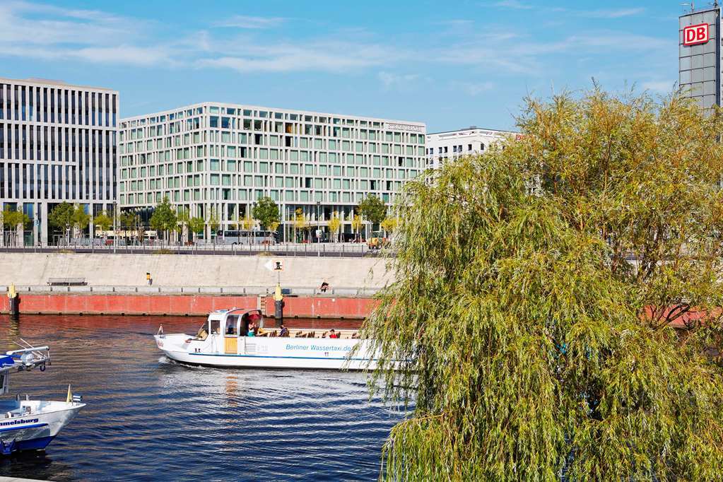 Steigenberger Hotel Am Kanzleramt Берлін Екстер'єр фото The photo shows a scenic view of a river with a boat traveling along it. On the left side, there are smaller boats, and on the right, a large white boat is visible, likely for tourism or leisure. In the background, there are modern buildings with a s