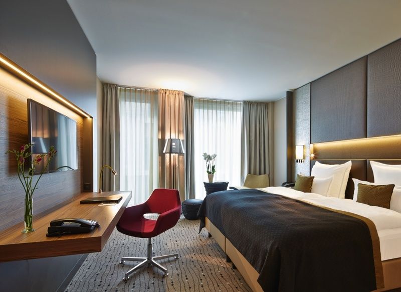Steigenberger Hotel Am Kanzleramt Берлін Екстер'єр фото The photo shows a modern hotel room with a contemporary design. It features a large bed with white pillows and a dark gray blanket. There is a sleek wooden desk and a red chair in the corner, along with a small black table. The room has large windows