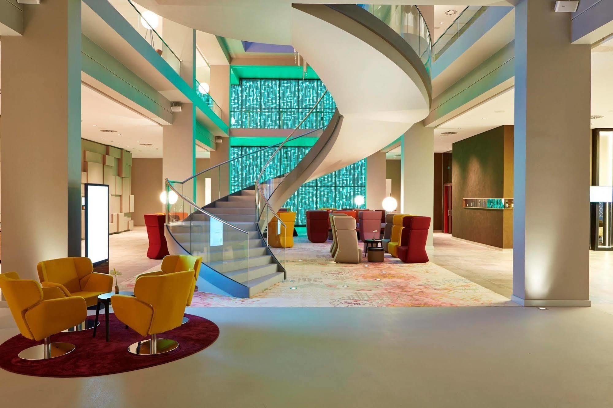 Steigenberger Hotel Am Kanzleramt Берлін Екстер'єр фото The photo shows a modern and stylish interior space, likely a hotel lobby. In the center, there's a prominent curved staircase that leads to an upper level. The walls are adorned with a colorful panel of glass or light, adding a vibrant touch to the 