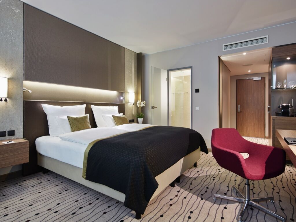 Steigenberger Hotel Am Kanzleramt Берлін Екстер'єр фото The image shows a modern hotel room featuring a neatly made bed with white linens and a dark quilt cover. On the bed, there are two decorative pillows. The walls are a soft gray color, and there is subtle lighting above the headboard. To the side, th