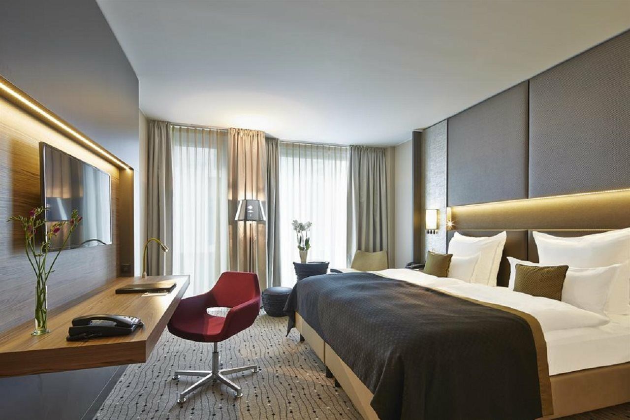 Steigenberger Hotel Am Kanzleramt Берлін Екстер'єр фото The photo shows a modern hotel room with a clean and stylish design. It features a large bed with plush pillows and a dark bedspread. There's a wooden desk with a chair, a lamp, and a decorative vase with flowers. Natural light filters in through lar
